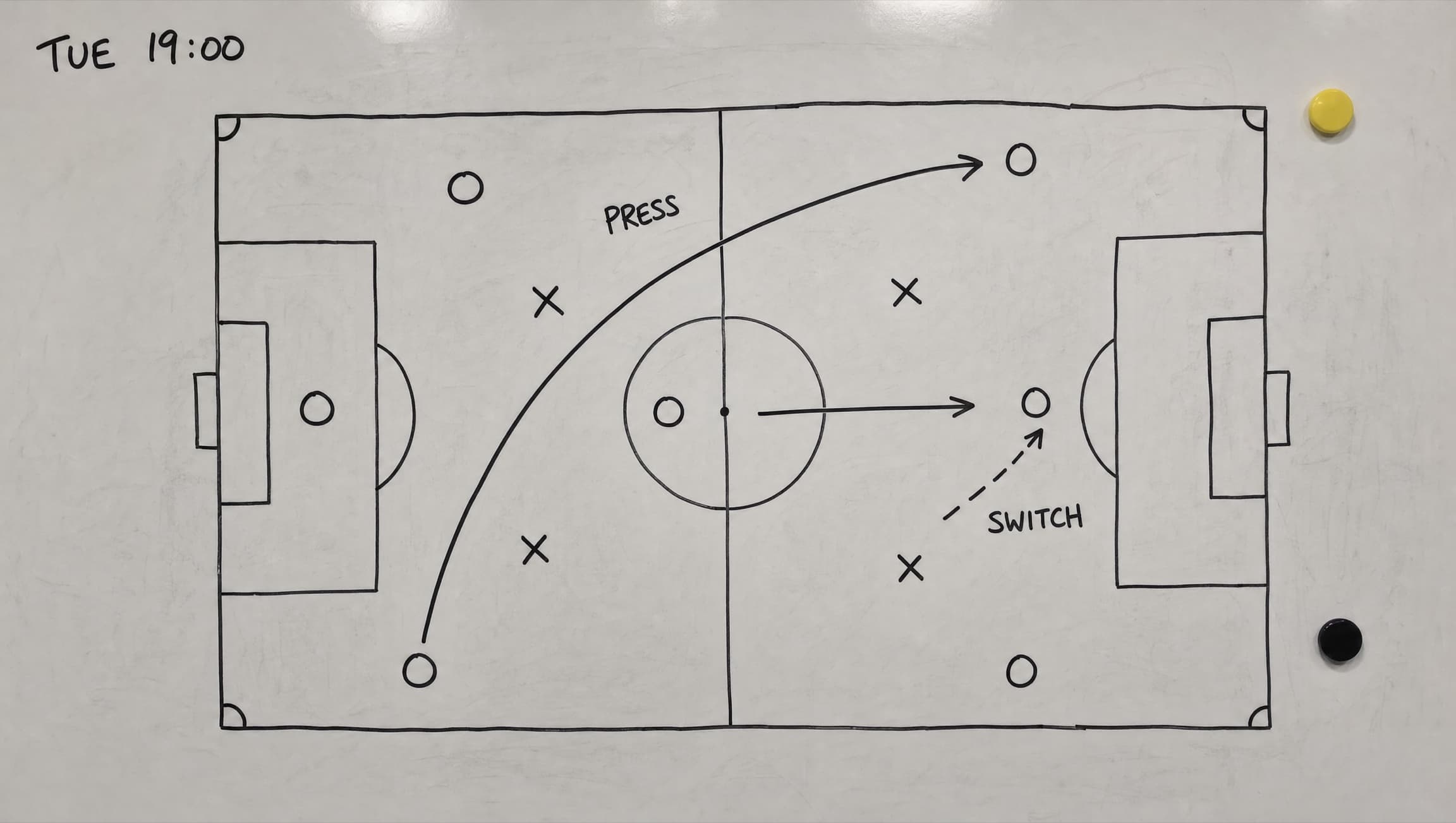The coach's training board: a hand-drawn marker pitch with O and X marks, a curved pressing arrow, a switch-of-play arrow, and the note TUE 19:00.